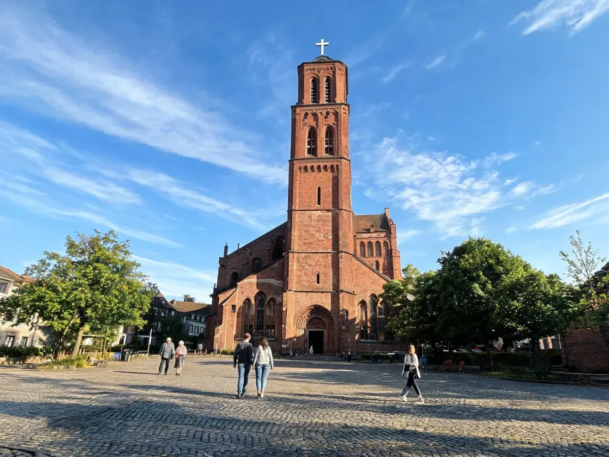 Wierni w słoneczne popołudnie przed zabytkowym kościołem Podwyższenia Krzyża Świętego w Poznaniu, spokojna atmosfera.